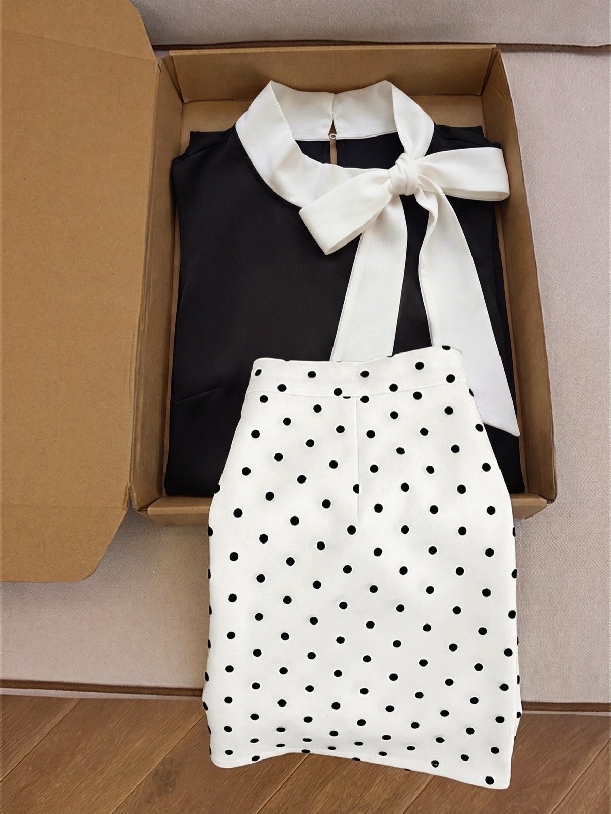 Boxed set flat-lay of a black bow-collar blouse and white polka-dot mini skirt set.